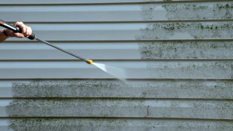 A man uses a power washer to clean vinyl siding on a home